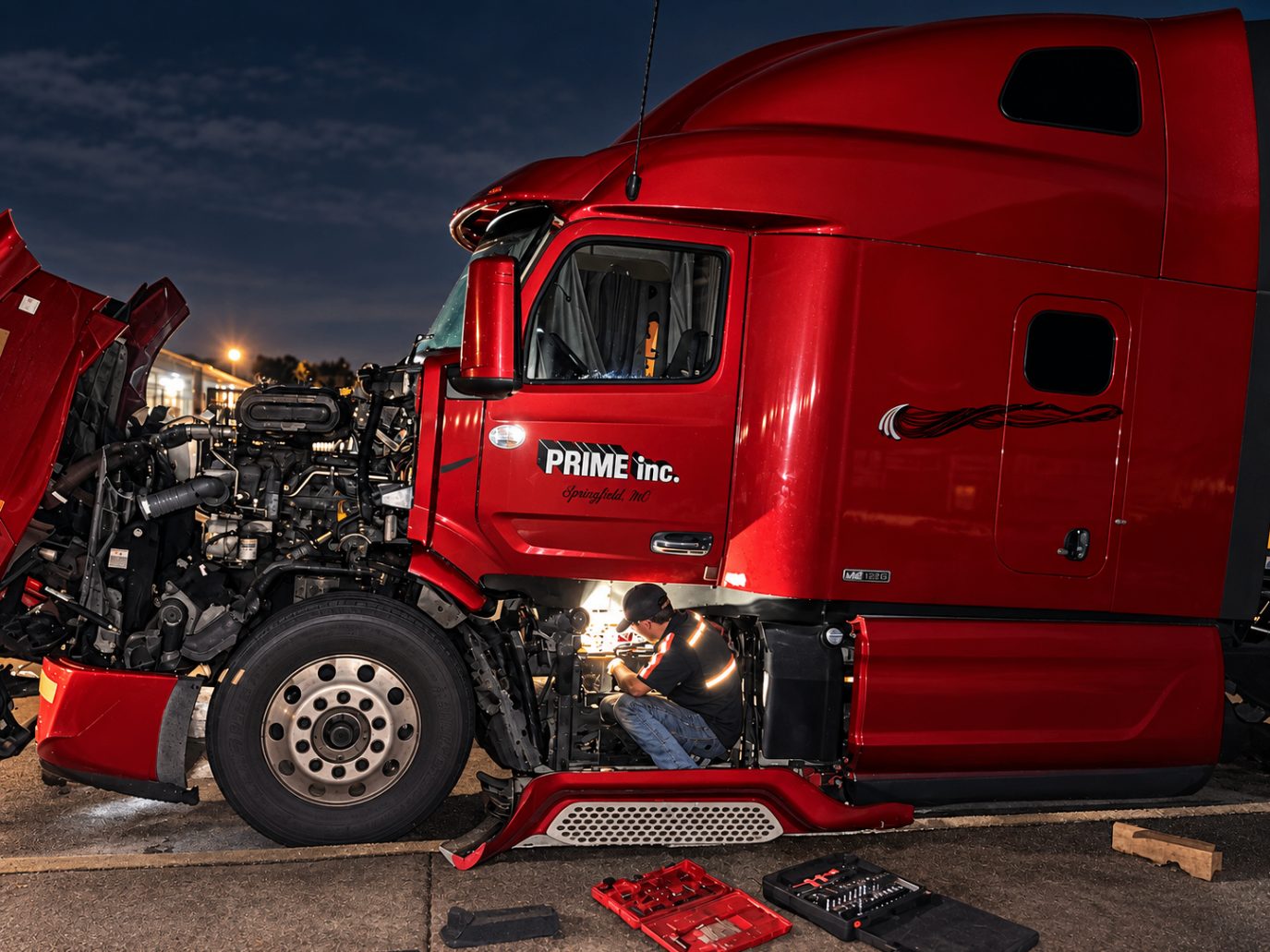 Justin working under the cab of a red semi truck at night with the hood tilted open