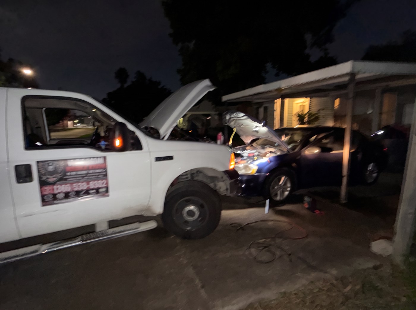 Freedom service truck and a car with raised hoods during a nighttime call beneath a carport